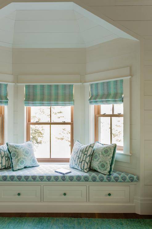 Turquoise blue roman shades hang from bay windows surrounded by white shiplap and located over a white built-in storage bench with turquoise blue knobs. The bench is topped with a turquoise blue and green cushion accented with turquoise blue and green pillows.