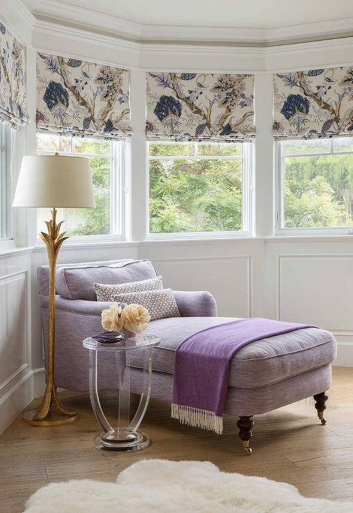 Purple chaise lounge in the corner of a master bedroom bay window beside a gold leaf floor lamp and a round lucite accent table. Bay window wainscoting adds a decorative touch to the walls with windows dressed with purple floral shades.
