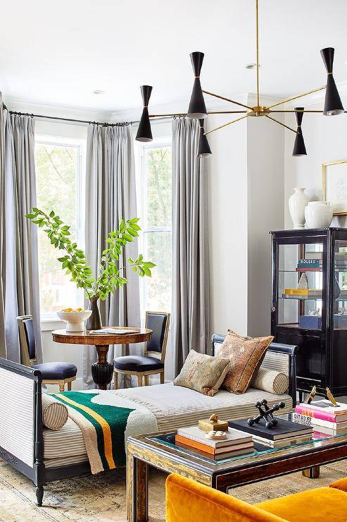 Contemporary living room designed with a black French daybed and stripe bolster pillows on a gray and tan rug in front of a bay window with black French chairs and an antique table.