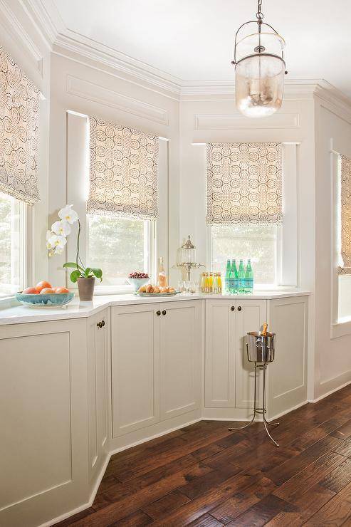 Kitchen bay window with tan shades features ivory built in cabinets accented with oval bronze knobs and white and silver marble countertops and dark wooden floors, illuminated by a mercury glass lantern.