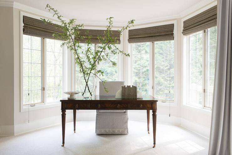 Transitional home office bay window designed with a French desk centered in the forefront paired with a slipcovered light gray chair. Brown bamboo roman shades finish the windows with a darker trim contrasting the natural linen desk chair.