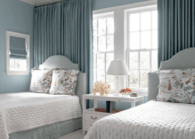Lovely blue shared bedroom features a shared white nightstand placed on a light gray rug beneath windows complemented with blue curtains hung behind light blue French headboards. The beds are finished with blue bed skirts and chinoiserie print pillows, and are lit by a Thomas O'Brien Bryant Chandelier.