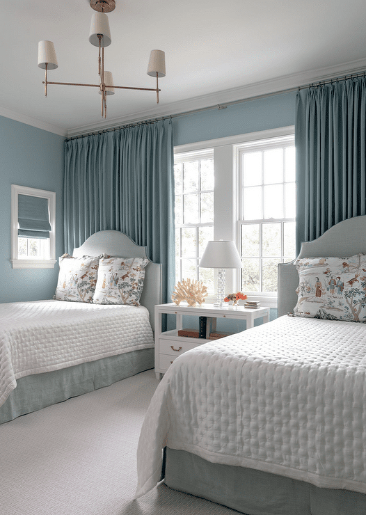 Lovely blue shared bedroom features a shared white nightstand placed on a light gray rug beneath windows complemented with blue curtains hung behind light blue French headboards. The beds are finished with blue bed skirts and chinoiserie print pillows, and are lit by a Thomas O'Brien Bryant Chandelier.