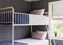 A red rope lantern lights a shared kid's bedroom boasting walls covered in black geometric wallpaper and a white metal bunk bed dressed in pink and blue bedding. A white rope and wool stool sits beside the beds on a tan striped rug.