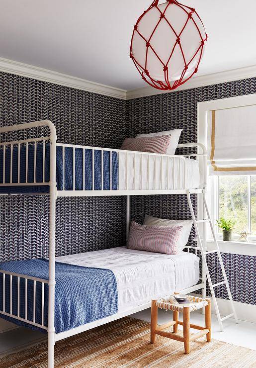 A red rope lantern lights a shared kid's bedroom boasting walls covered in black geometric wallpaper and a white metal bunk bed dressed in pink and blue bedding. A white rope and wool stool sits beside the beds on a tan striped rug.