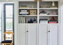 An antique brass picture light is fixed beneath a white plank ceiling and over white built-in bookshelves, styled and fitted over white shaker built-in cabinets donning oval knobs.