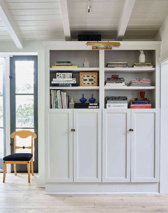 An antique brass picture light is fixed beneath a white plank ceiling and over white built-in bookshelves, styled and fitted over white shaker built-in cabinets donning oval knobs.