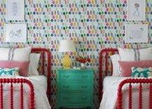 Colorful wallpaper complements a charming kids' bedroom featuring red spindle beds placed beneath stacked art pieces and flanking a yellow lamp topping a green French nightstand.