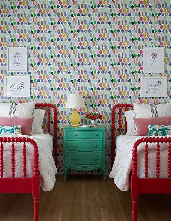 Colorful wallpaper complements a charming kids' bedroom featuring red spindle beds placed beneath stacked art pieces and flanking a yellow lamp topping a green French nightstand.