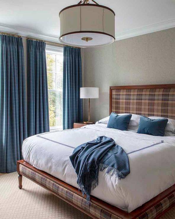 Welcoming blue and brown bedroom features a brown plaid bed accented with blue velvet lumbar pillows and a blue throw placed on blue hotel bedding. The bed is positioned on a tan diamond pattern rug and in front of a gray wallpapered wall, as blue curtains cover windows.