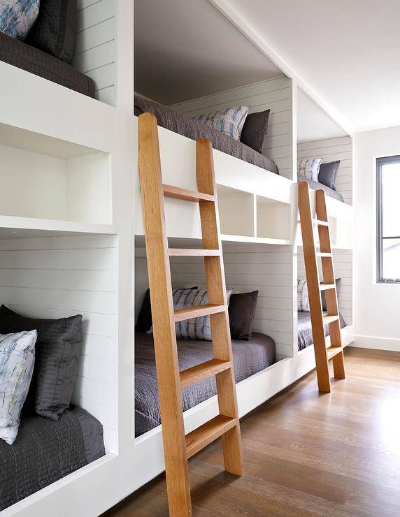 Bunk room for 6 features white custom built-in bunk beds fixed in a row and accented with built in shelves, shiplap trim, black bedding, and brown wooden ladders.