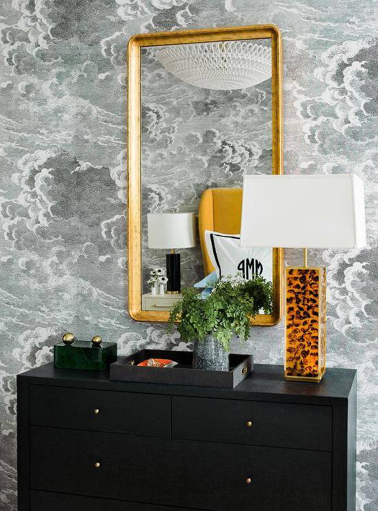 Fornasetti Nuvole Wallpaper complements a black dresser styled with a black tray and a tiger lamp and placed beneath a curved brass mirror.