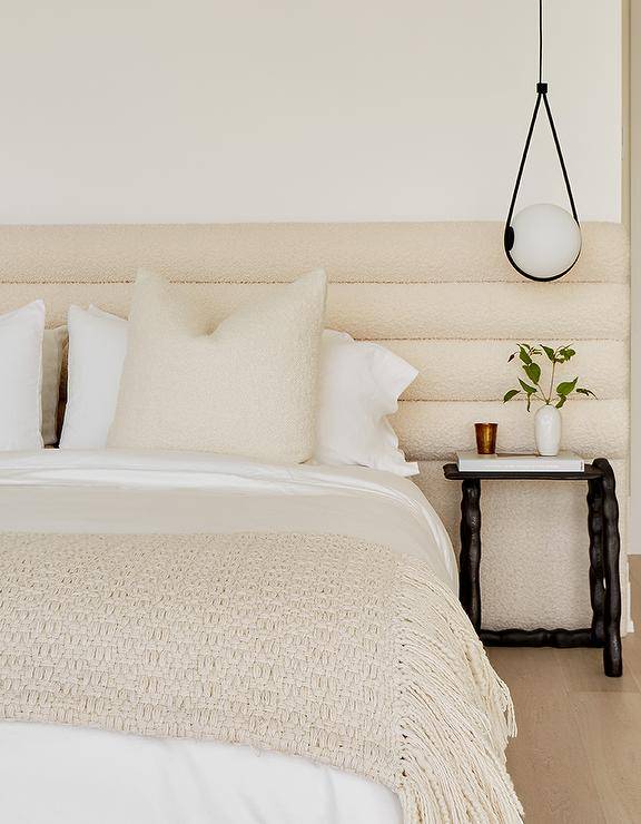 The bedroom features a cream boucle channel tufted headboard accented with cream boucle pillows, white and cream bedding, cream knit throws, and black sawtooth bedside tables. .