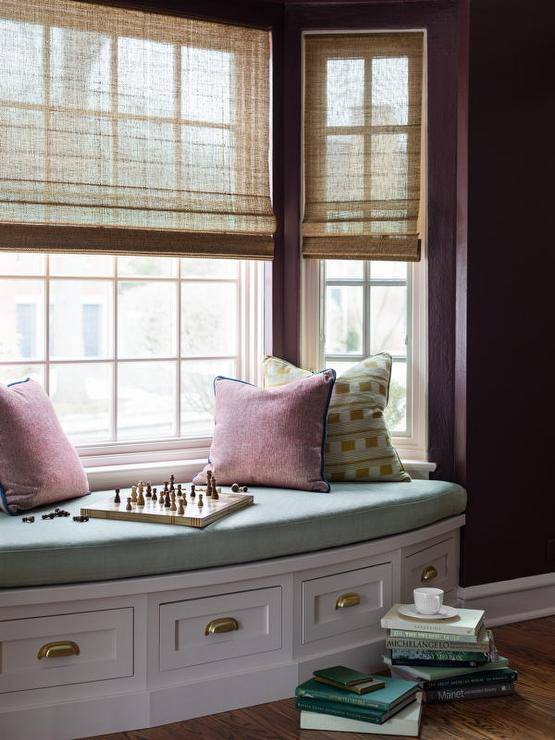 Bay windows covered in bamboo roman shades are located over a white built-in window seat boasting drawers with brass cup pulls and a blue velvet cushion topped with red and green pillows.