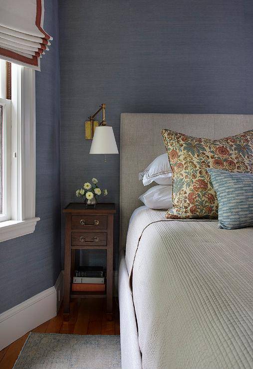 Bedroom features a light gray tufted bed on blue grasscloth wallpaper and a small tall wooden nightstand lit by a brass swingarm sconce.