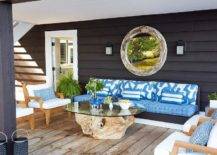 Stunning covered patio features a round driftwood mirror hung from black home siding over a black and white sofa accented with blue cushions topped with white and blue striped pillows. Gold teak chairs sit on teak floor planks facing a glass and driftwood coffee table.
