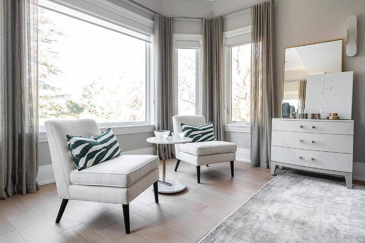 Sheer gray curtains cover bay windows framed by light gray wall paint and positioned behind a nickel and marble accent table placed between white slipper chairs topped with green zebra pillows.
