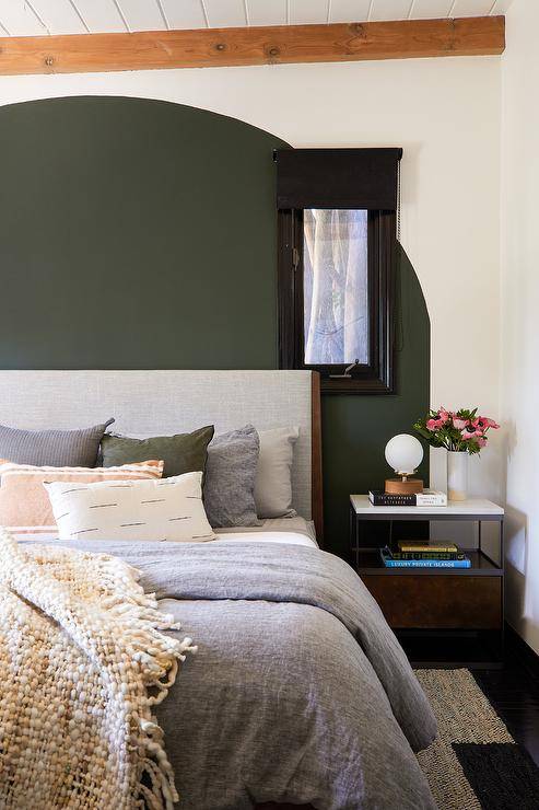 An arched under green painted accent wall is positioned behind a vintage gray fabric and wooden headboard accenting a bed dressed in gray bedding topped with gray, green and tan pillows. A metal and marble nightstand is positioned beside the bed and beneath a window.