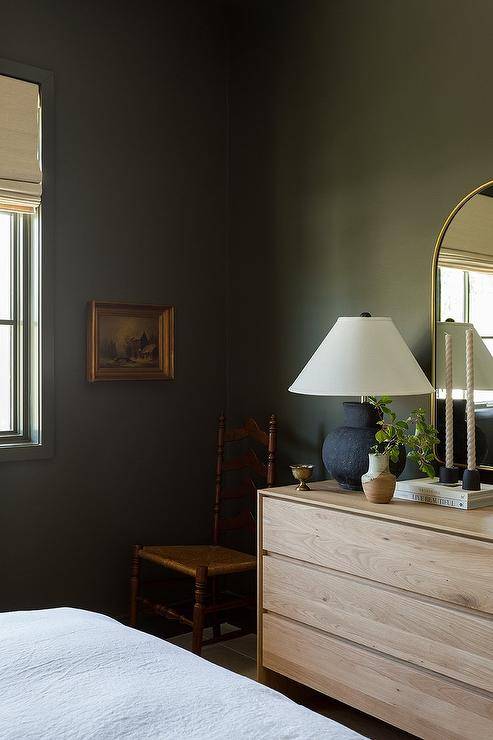 A light beige dresser with brass arch mirror lit by a black vintage lamp, sits on a moss green painted wall with wooden chair.