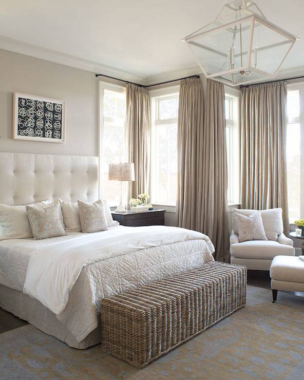 Elegant bedroom with black and white framed art on greige walls, bed with ivory tufted headboard and ivory and white bed linen. A black nightstand sits next to the bed, and a white table lamp sits next to a wall of windows with floor-length taupe curtains. A white lantern pendant hangs at the foot of the bed, and a wicker bench rests on a gold and blue area rug.
