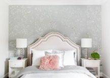 Sophisticated bedroom features a tan French headboard that supports a bed with white bedding and a pink faux fur pillow, flanked by glass and nickel lamps over white nightstands on an accent wall clad in gray floral wallpaper.