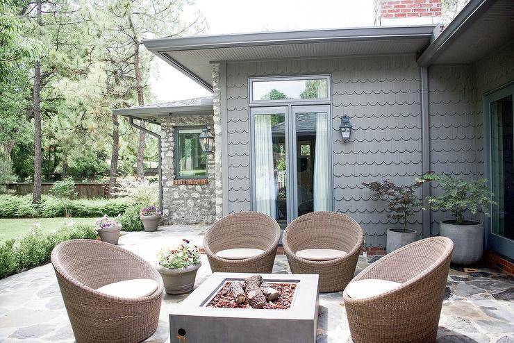 Wicker pod chairs surround a rectangular concrete fire pit placed in front of a home clad in gray scalloped siding.
