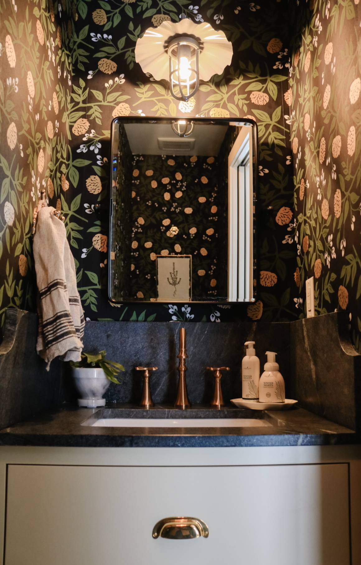 dark floral wallpaper on walls and ceiling surrounding dark bathroom vanity with black mirror