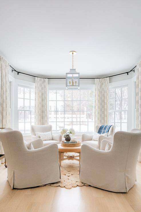 A white pagoda lantern hangs over a round brown leather ottoman placed on a crochet rug ad surrounded by white wingback lounge chairs. The chairs sit in front of bay windows dressed in ivory and black curtains.