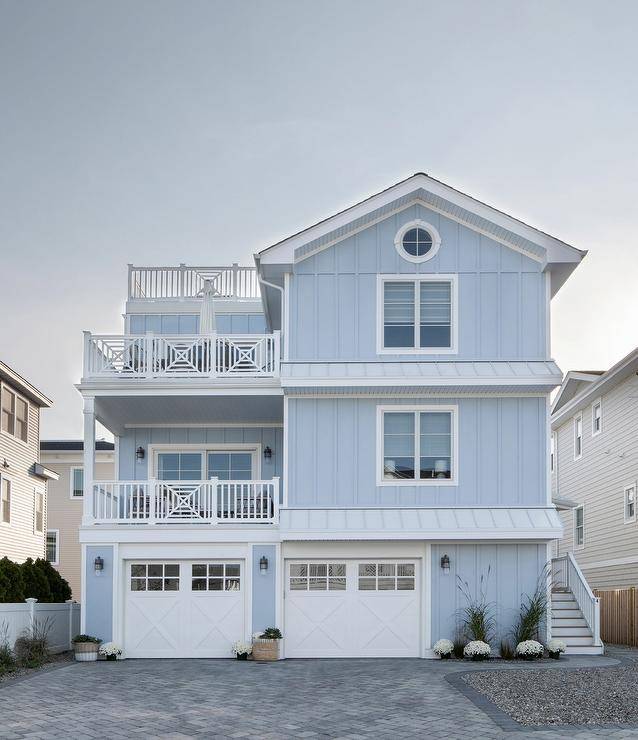 Periwinkle beach house boasting white trim with two white x-trimmed balconies, vertical siding, white double garage doors and gray pavers.