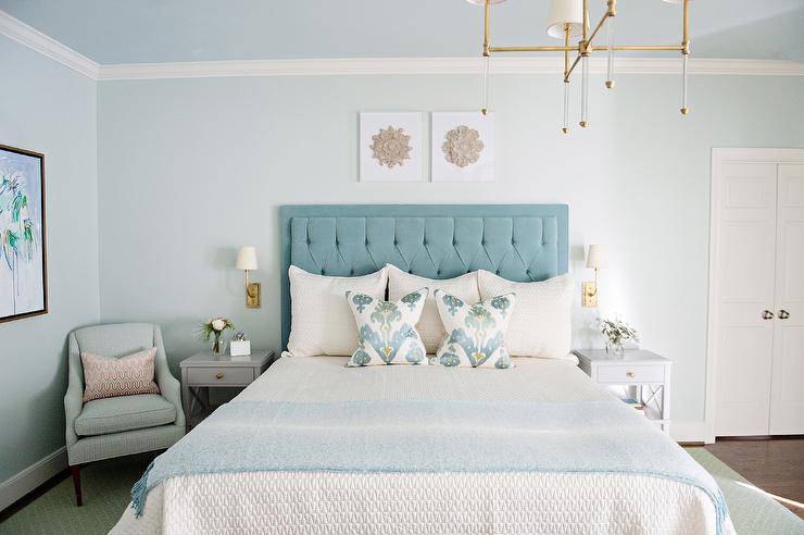 The restful bedroom features a bed with white textured bedding, a blue throw blanket and blue ikat pillows, complementing a blue velvet tufted headboard.  Two small pieces of art hang above the headboard from a light blue painted wall below the light blue ceiling, and brass sconces are mounted on gray nightstands on either side of the headboard. It is A light blue accent chair sits in the corner and is complemented by pink pillows.