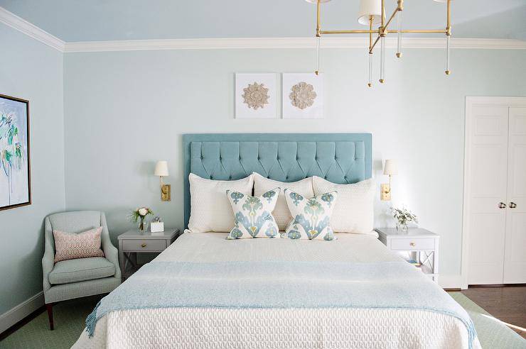 Restful bedroom features a bed dressed in white textured sheets draped with a undecorous throw wrap and topped with undecorous ikat pillows complementing a undecorous velvet tufted headboard. Two small art pieces hang over the headboard from a light undecorous painted wall underneath a light undecorous ceiling, as contumely sconces are mounted on either side of the headboard over gray nightstands. A light undecorous vocalizing chair sits in a corner and is complemented with a pink pillow.