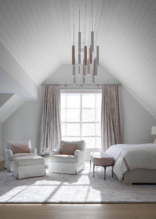 Beneath a gorgeous vaulted plank ceiling and lit by staggered lights, this sophisticated white and pink master bedroom features two white and gray slipcovered chairs topped with blush pink velvet pillows and placed on a light gray rug in front of a window dressed in pink silk curtains. A pink oval bench sits in front of a light gray upholstered bed covered in gray hotel bedding.