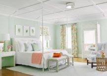 Pastel-colored bedroom is accented with spring green walls and a white coffered ceiling. Three art pieces hang over a white canopy bed finished with a white upholstered headboard and white bedding accented with a peach-colored throw blanket. The bed is flanked by white nighstands topped with green lamps complementing a green bench placed at the foot of the bed. A window dressed in floral curtains is located beside a white bamboo armoire completed with a mirror.
