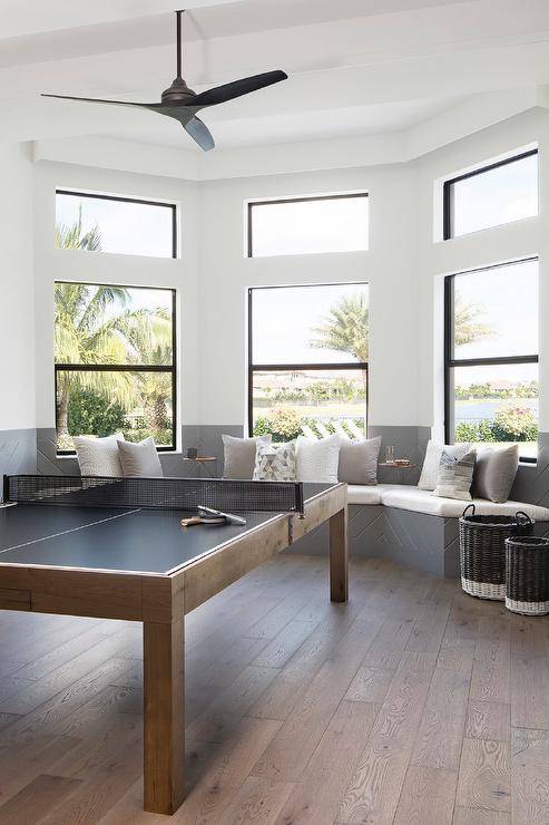 Chic game room features a bay window fitted with gray window seats alongside white and gray cushions. A modern ping pong sits in front of a bay window.