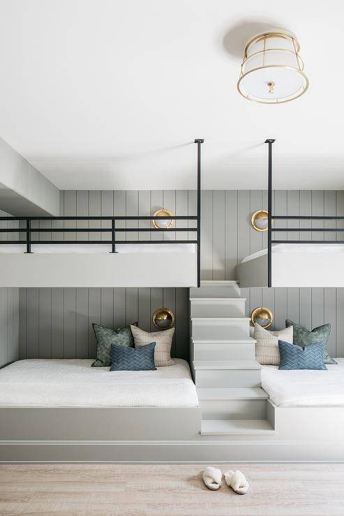A pair of light gray bunk beds are separated by a light gray staircase, fixed against gray vertical plank trim, and accented with black railing lit by brass porthole sconces.