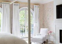 A white faux bois chandelier lights a bedroom clad in pink chinoiserie wallpaper and boasting a light gray canopy bed placed on a light gray rug. Tall arched windows are covered in white curtains hung from gold rods, while a white corner chair and ottoman sit in a corner beside a white spindle accent table.