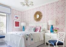 Contemporary bedroom features a white tufted headboard accented with pink pillows flanked by white nightstands lit by Caribbean blue lamps, a white chair with blue cushion and a gold sunburst mirror mounted on Brunschwig & Fils les touches pink wallpaper.