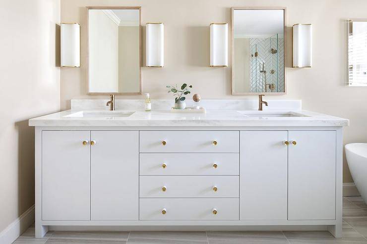 White and gold bathroom boasts a white dual washstand accented with polished pulls and a white marble countertop. Gold polished brass vintage cross handle faucets complement the warmer tones in this light bathroom. Two thin wood framed vanity mirrors are illuminated by a a set of white glass and brass sconces. A light neutral beige painted wall adds a sense of warmth to the bathroom allowing the decor to emphasize minimalism.