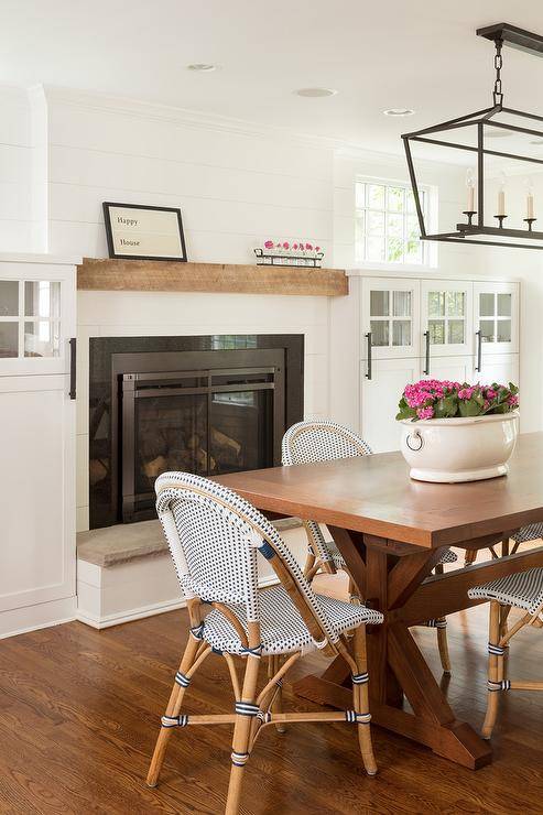 Darlana Linear lantern illuminates a trestle dining table with navy blue French bistro chairs in a cottage home design featuring a fireplace and hearth.