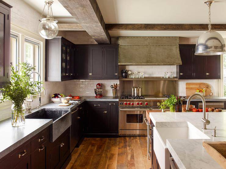 Cottage style kitchen features vintage latch hardware on black shaker cabinets topped in concrete, a French kitchen hood over a stainless steel cooktop backsplash with stainless steel shelf and a wolf dual range, glazed subway tiles, a marble topped island with sink that boasts a nickel gooseneck faucet lit by a nickel dome lantern, dark wooden floors and wooden ceiling beams.