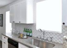 Well appointed white kitchen features a stainless steel dishwasher fixed between white shaker cabinets accented with oil rubbed bronze knobs and a gray quartz countertop finished with a stainless steel curved sink and a polished nickel hook and spout faucet. The faucet is mounted beneath a window lit by an accordion style sconce and partially framed by Home Depot Merola Tiles.