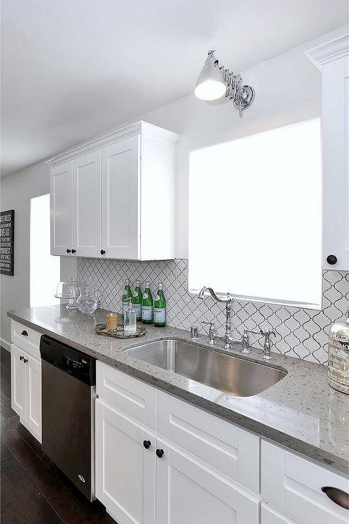 Well appointed white kitchen features a stainless steel dishwasher fixed between white shaker cabinets accented with oil rubbed bronze knobs and a gray quartz countertop finished with a stainless steel curved sink and a polished nickel hook and spout faucet. The faucet is mounted beneath a window lit by an accordion style sconce and partially framed by Home Depot Merola Tiles.