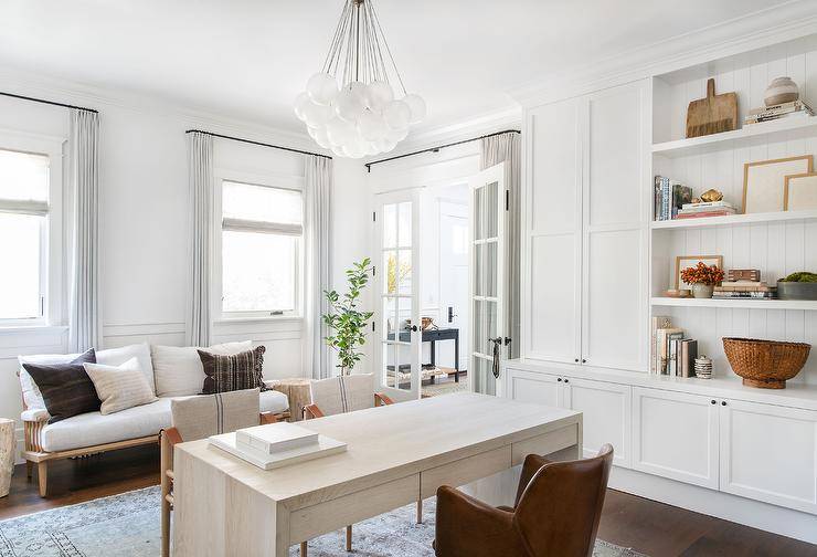 A brown leather desk chair sits at a lime oak desk facing two French burlap and leather chairs placed on a gray vintage rug and lit by a white glass bubbles chandelier. White built-in shelves are mounted against a white plank backsplash and over white shaker cabinets with oil rubbed bronze knobs.