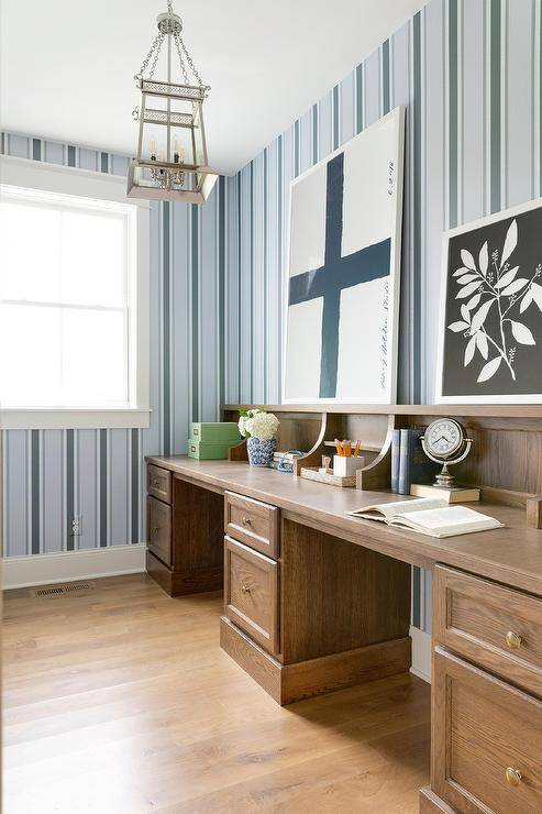 Cottage style home office with blue and black vertical stripe wallpaper, a brown oak vintage style desk and antique brass knobs. Wall art and desk decor bring a cozy and sophisticated appeal in this cottage office space.