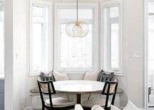 A bay window mini breakfast nook features a round marble and brass dining table with black cane dining chairs and a bay window built in bench near a white sofa.