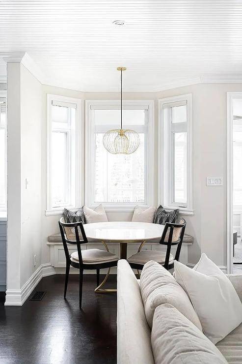 A bay window mini breakfast nook features a round marble and brass dining table with black cane dining chairs and a bay window built in bench near a white sofa.