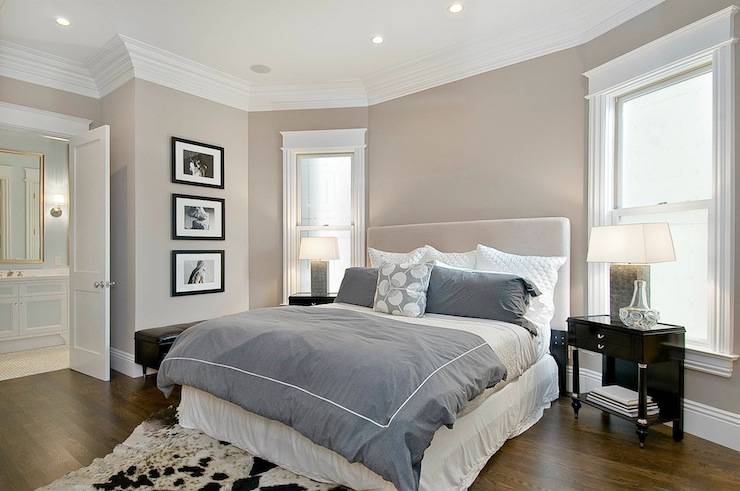 Super master bedroom with greige walls paint color, light gray headboard, blue purple duvet & shams, white ruffled bedskirt, black & white cowhide rug, espresso stained glossy nightstands and crown molding.