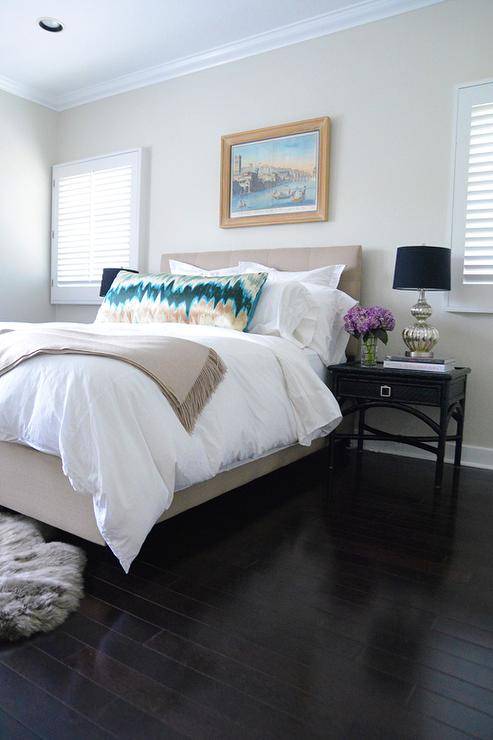 Between square windows covered in shades, an oil painting hangs over a beige upholstered bed dressed in white hotel bedding topped with a gold and teal body pillow and a camel colored throw blanket. The bed is flanked by black bamboo nightstands topped with silver fluted lamps accented with donning lamp shades lighting dark wood floors in this restful beige and black bedroom.