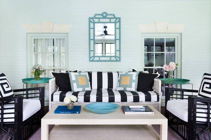 Gorgeous sunroom features a beadboard sloped ceiling accented with a ceiling fan cooling a blue bamboo mirror, Bungalow 5 Chloe Mirror, placed over a white wicker sofa adorned with black and white cushions flanked by teal tray tables facing a Serena & Lily Blake Raffia Rectangular Coffee Table.