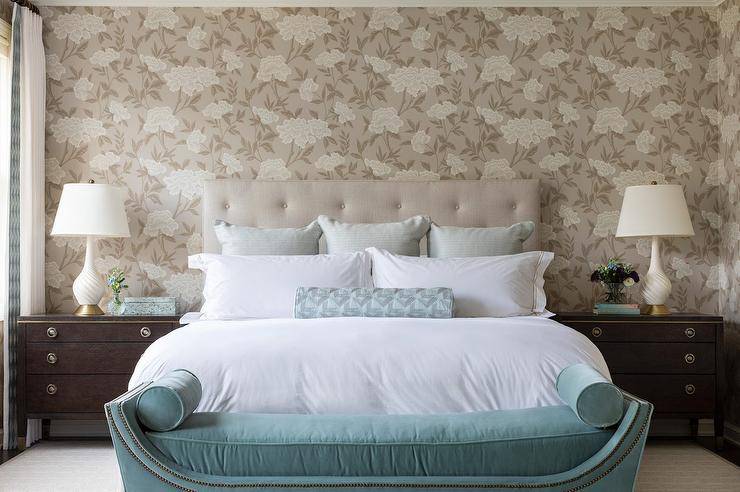 A light beige tufted headboard is fitted in front of a white and brown floral wallpapered wall and accents a bed complemented with a light blue bolster pillow. At the foot of the bed, a teal velvet bench topped with teal velvet lumbar pillows sits on a beige rug, as brown antique nightstands lit by ivory twist lamps flank the bed.
